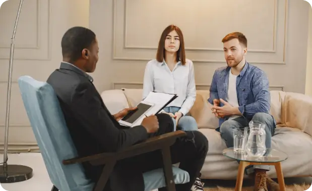 A male marriage counselor in Kailua, Oahu holds a tablet and clipboard while a young couple shares their experiences across a glass coffee table at a Windward Oahu therapy practice.